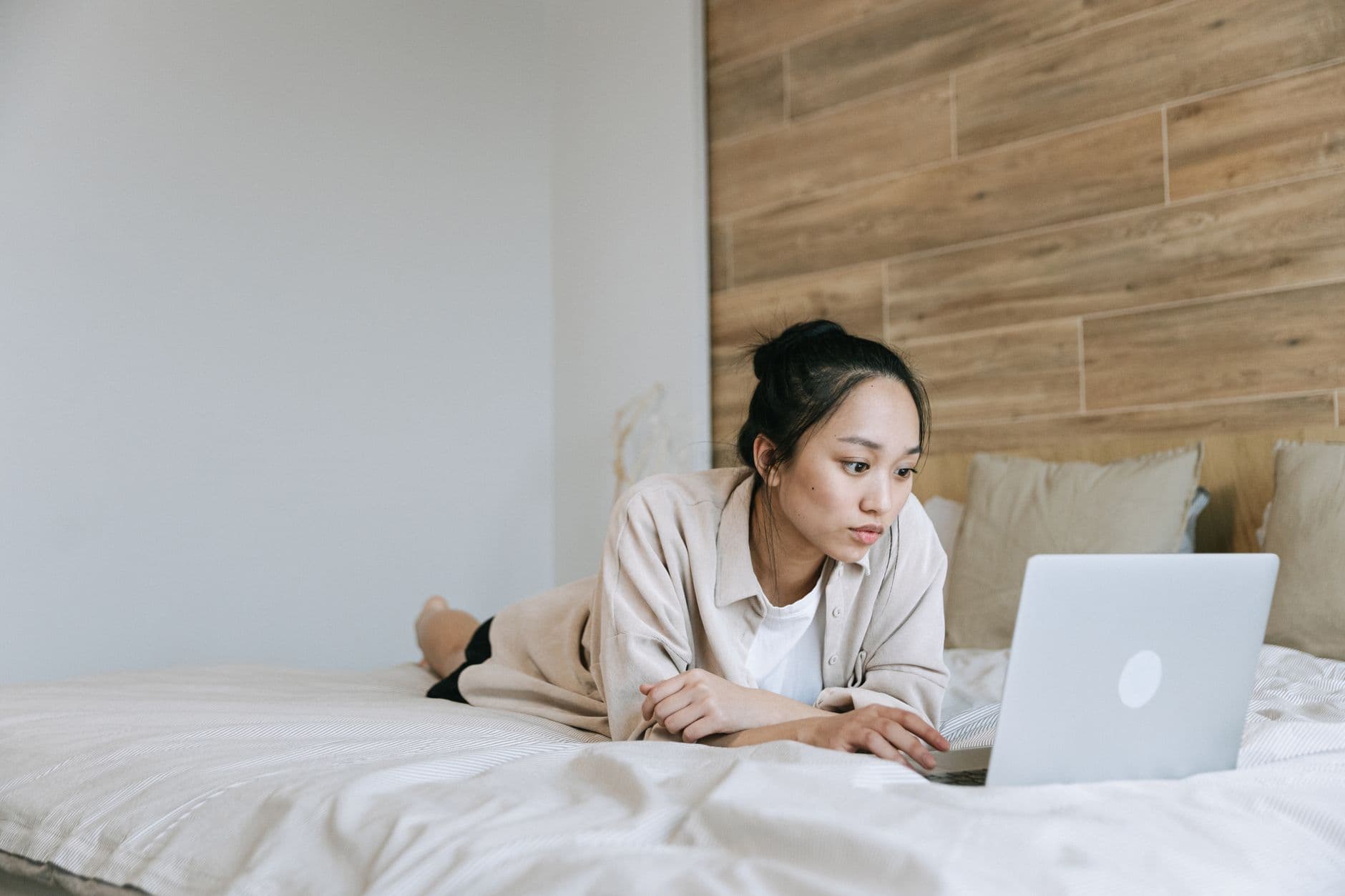 A woman with her hair in a bun lying on her bed, still focused on her laptop