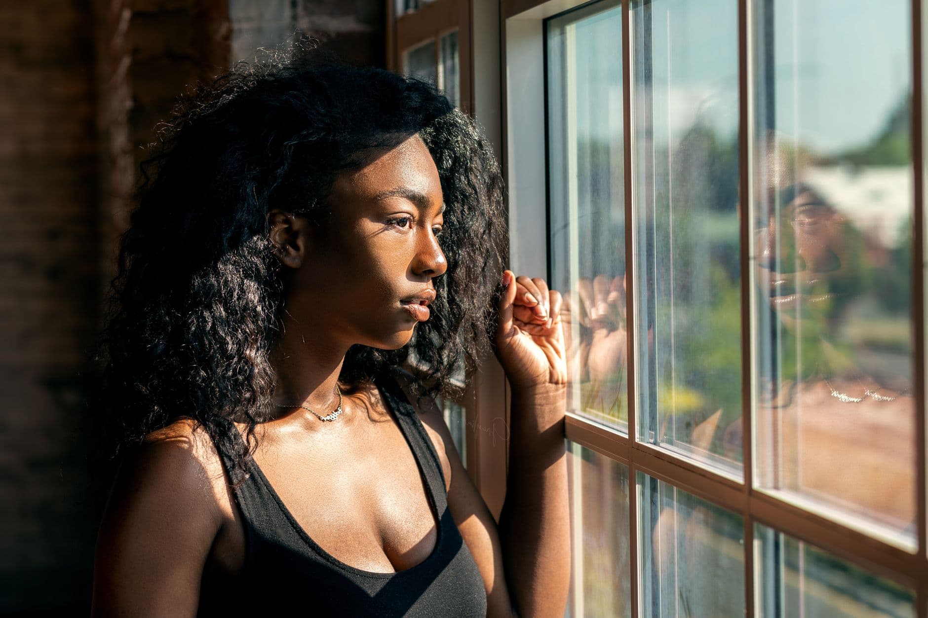 A Black woman standing at a sunlit window, looking out thoughtfully, holding the weight of how far she has come