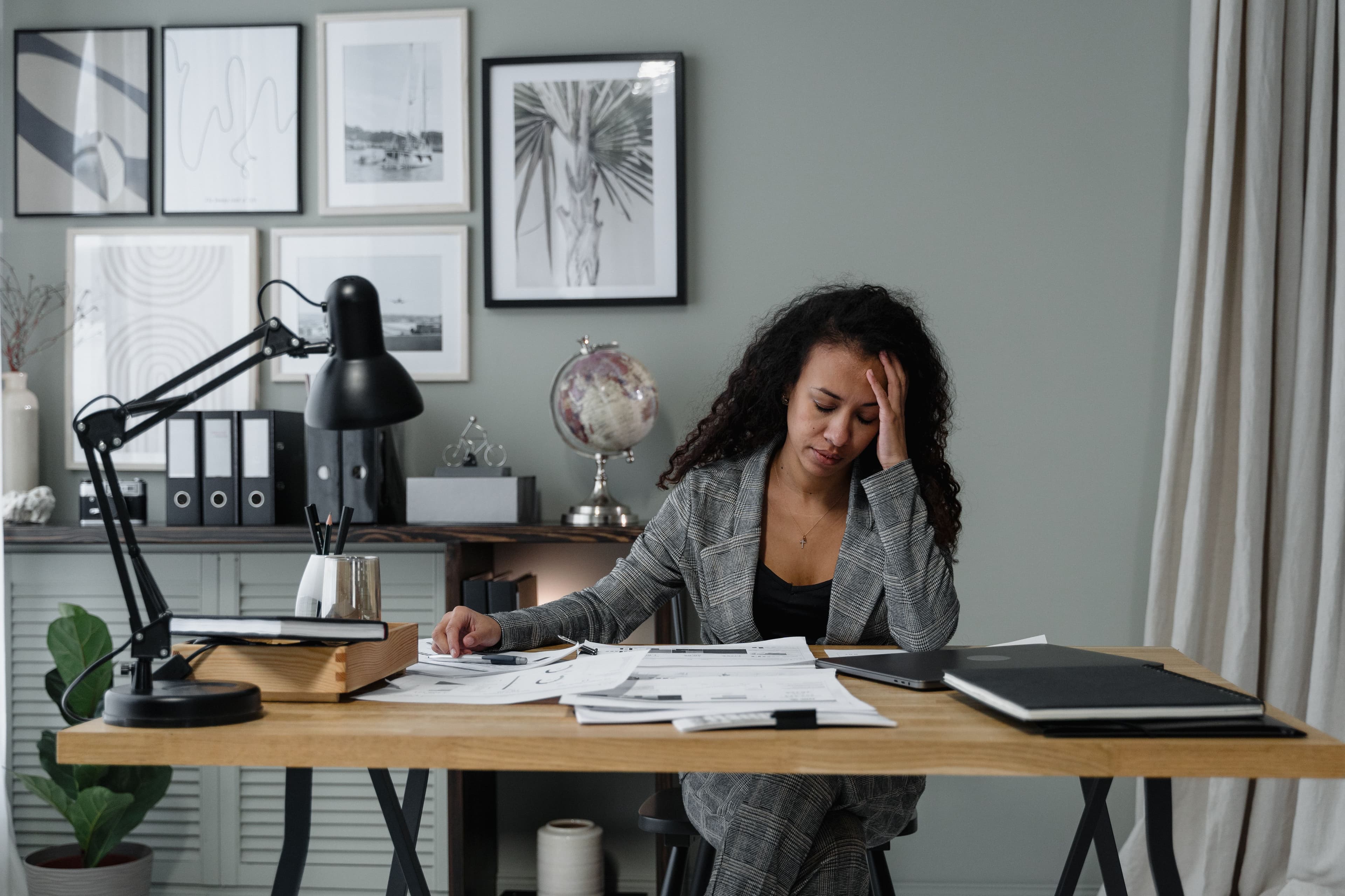 A woman of color at her desk surrounded by papers, hand pressed to her temple, capturing the quiet weight of high-functioning anxiety