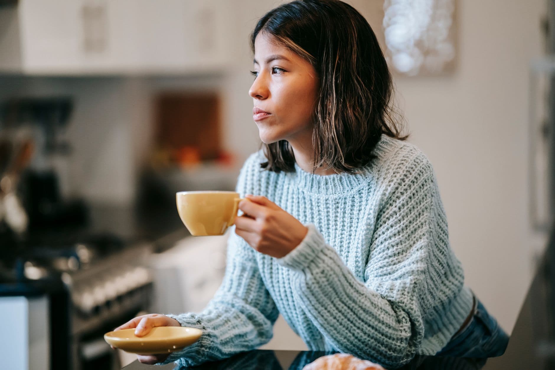 A woman in a soft knit sweater pausing with a cup of coffee in her kitchen, taking a quiet moment before her day starts