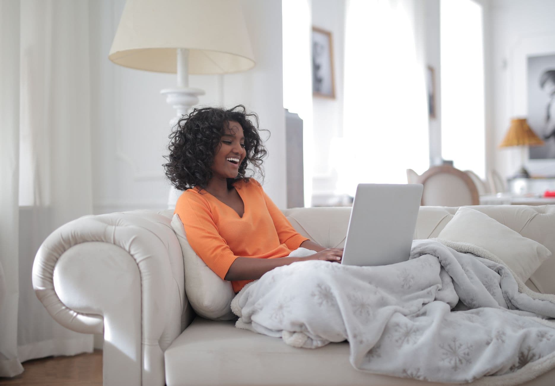 A Black woman laughing warmly while sitting on her couch searching on her laptop, natural light in a cozy home
