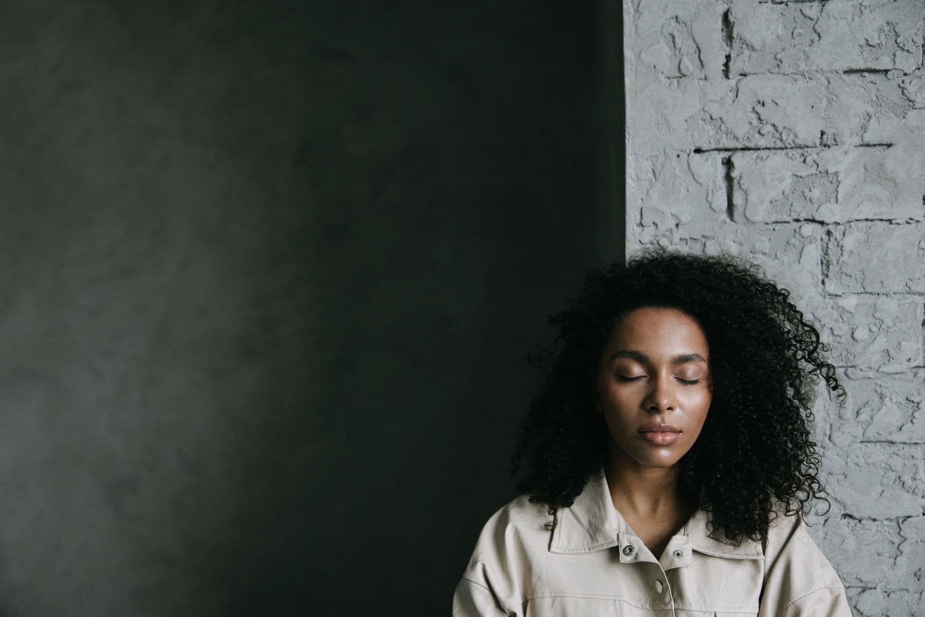 A woman with curly hair sitting with her eyes closed against a textured wall, breathing slowly