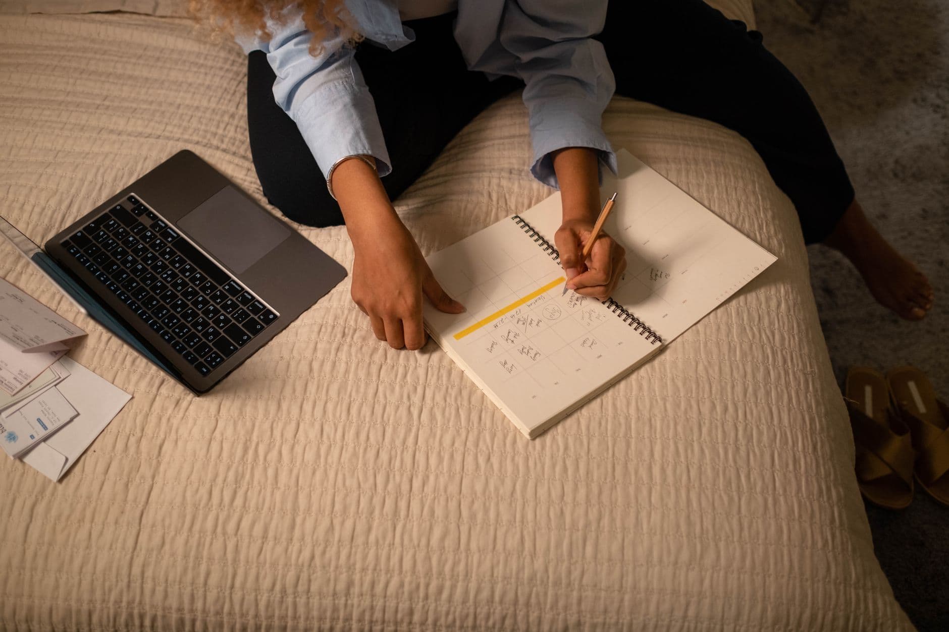 A woman writing plans in a spiral planner on her bed beside a laptop, warm evening light