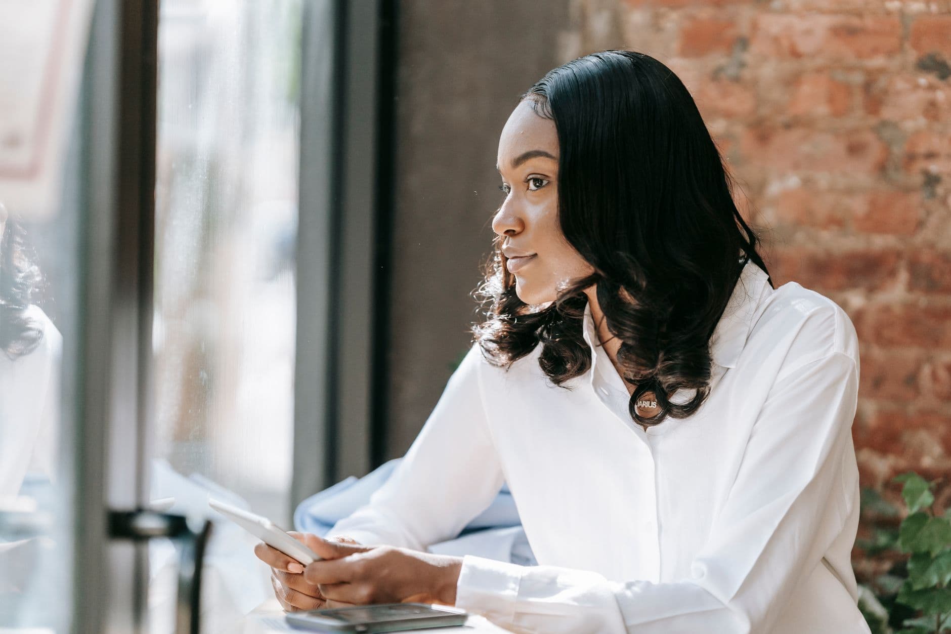 A Black woman in a white shirt sitting by a window, looking out, her phone in her hands